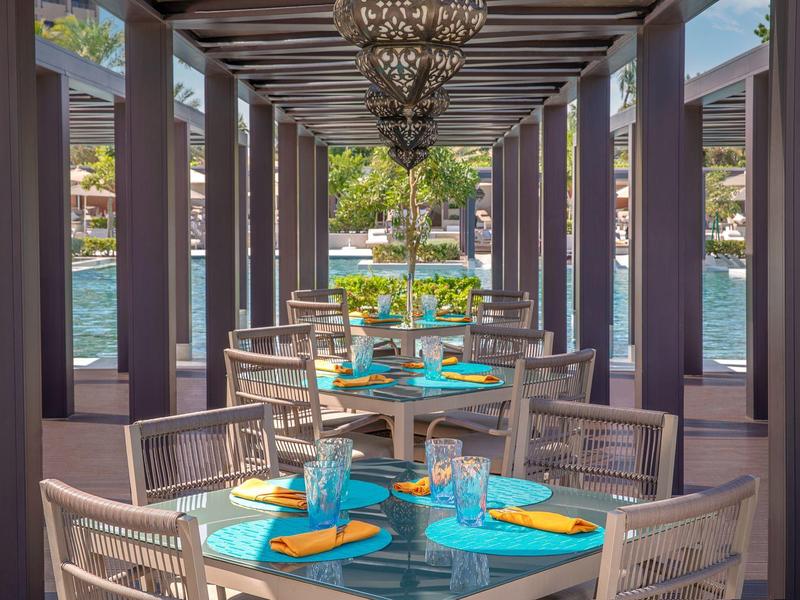 Jídelní prostor s modrými stoly a židlemi pod pergolou vedle výhledu na vodu.