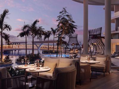 Elegantní restaurace s venkovním posezením, výhled na palmy a přístav při západu slunce.