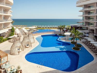 Moderní venkovní bazén s lehátky a výhledem na moře mezi dvěma hotelovými budovami.