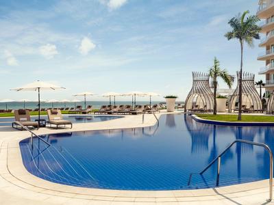Runder Hotelpool mit Liegestühlen und Sonnenschirmen, Palmen und Blick aufs Meer.