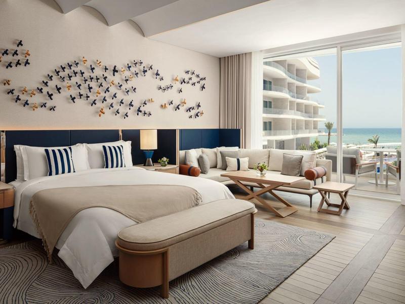 Moderní hotelový pokoj s velkou postelí, sedací částí a výhledem na moře z balkonu.