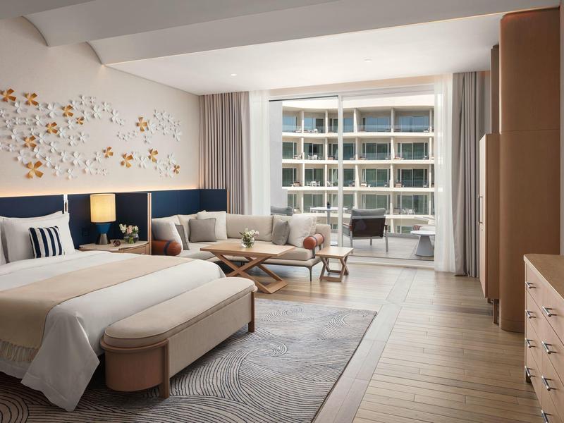 Moderní hotelový pokoj s manželskou postelí, posezením a balkonem s výhledem na sousední budovu.