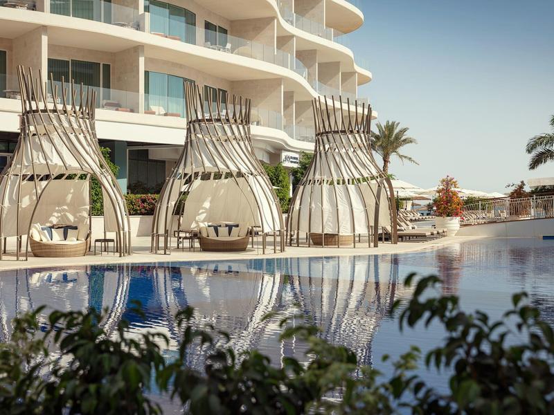 Moderní závěsné křesla a bazén před hotelem s oblými balkony při západu slunce.