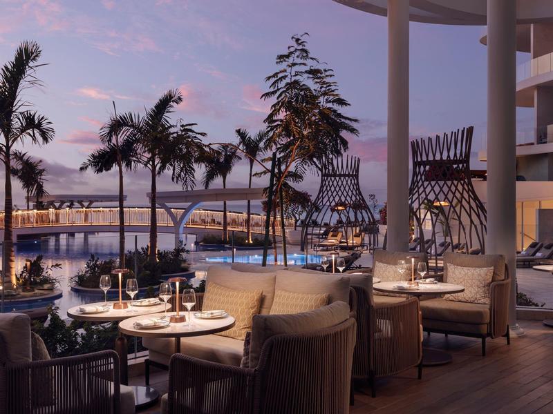 Elegantní restaurace s venkovním posezením, výhled na palmy a přístav při západu slunce.