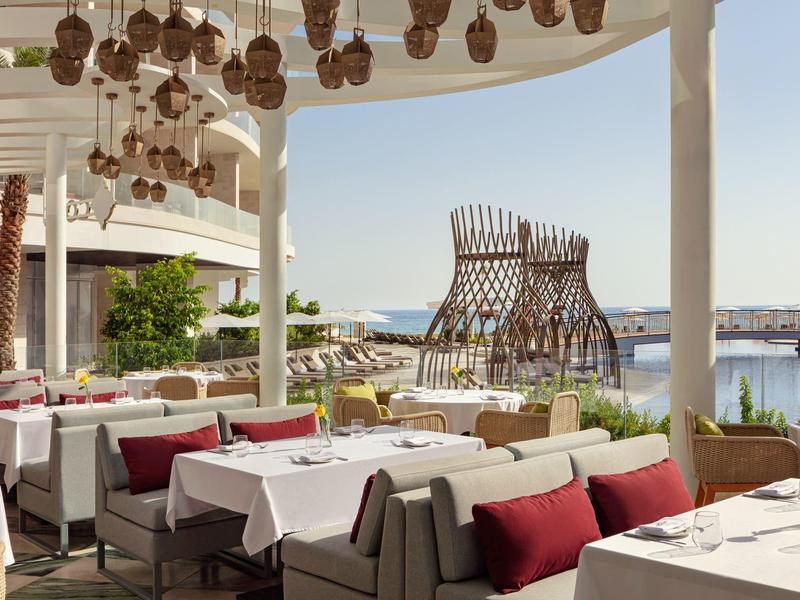 Otevřená restaurace s bílými stoly, červenými polštáři, dekorativními lampami a výhledem na přístav.