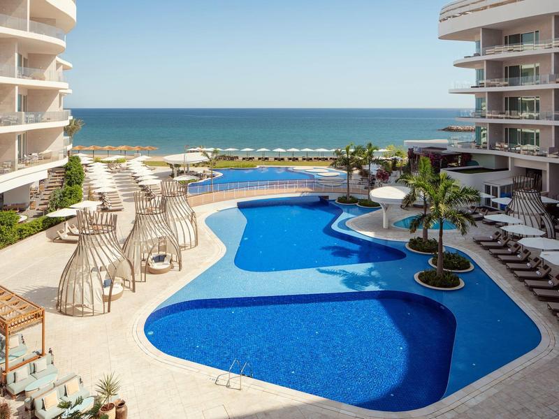 Moderní venkovní bazén s lehátky a výhledem na moře mezi dvěma hotelovými budovami.