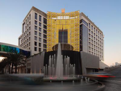 Hohes Gebäude mit gelb beleuchtetem Eingang und großer runder Fontäne davor bei klarem Himmel.