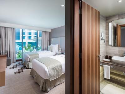 Modernes Hotelzimmer mit zwei Betten, großem Fenster, Vorhängen und angrenzendem Badezimmer.