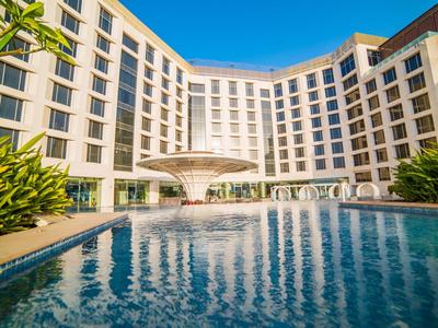 Modernes Hotelgebäude mit großem rechteckigem Pool und zentralem Wasserspiel bei klarem Himmel.