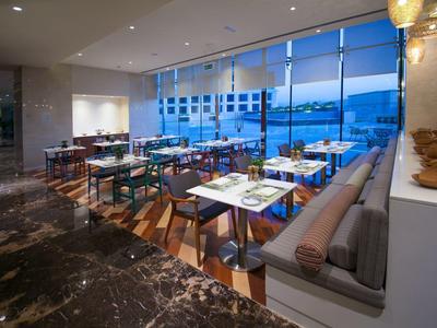 Moderner Speiseraum mit großen Fenstern, Holzstühlen, Tischen und Sofabank in neutralen Farben.