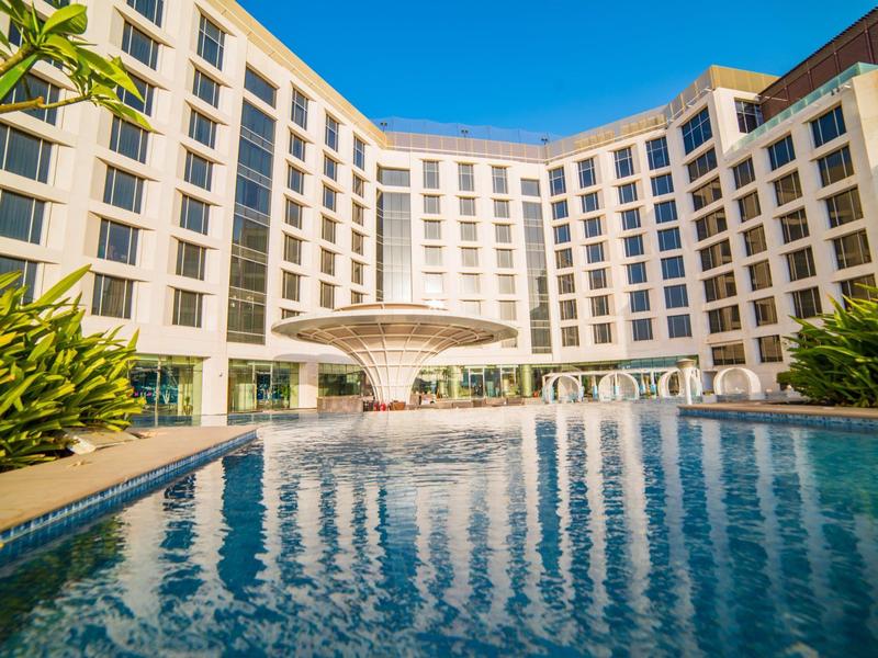 Modernes Hotelgebäude mit großem rechteckigem Pool und zentralem Wasserspiel bei klarem Himmel.