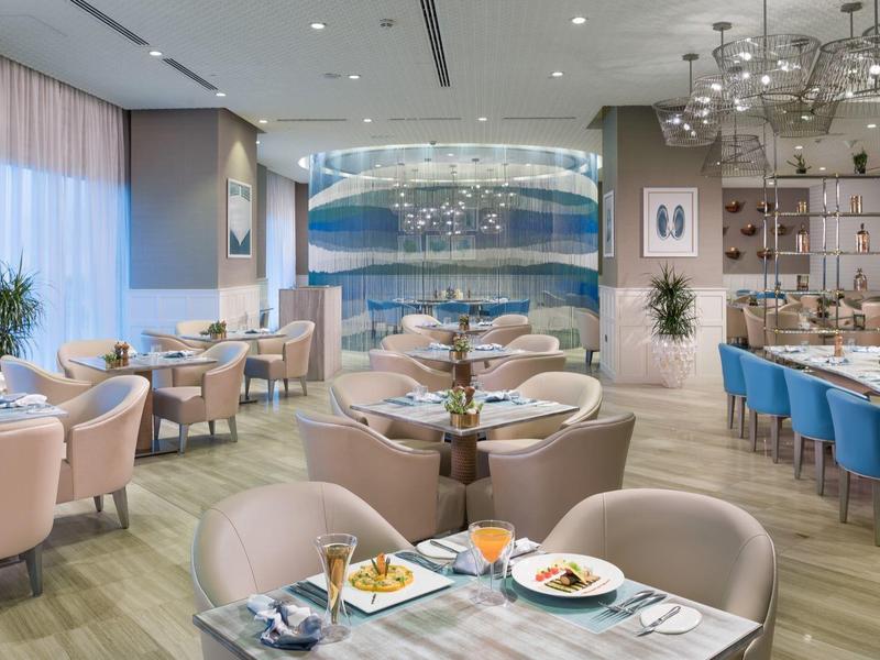 Moderner Restaurantraum mit rosafarbenen Sesseln, blauen Barstühlen, hängenden Lampen und großen Fenstern.