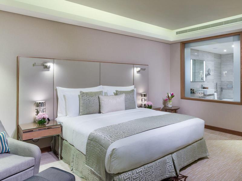 Helles Schlafzimmer mit großem Bett, grauem Kopfteil, zwei Nachttischen und Fenster zum Badezimmer.