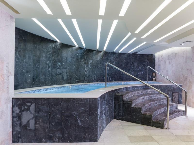 Moderner Raum mit Marmor-Treppen und eingetauchtem Pool, beleuchtet durch lange, helle Deckenleuchten.