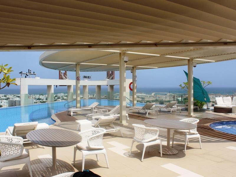 Terrassenbereich mit weißen Tischen und Stühlen, Pool, Glasgeländer und Blick aufs Meer.