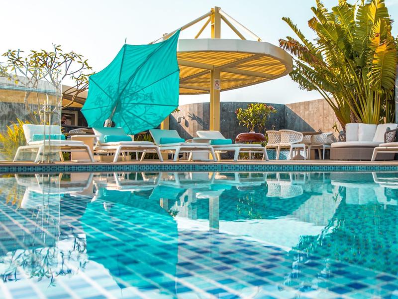 Schwimmbad mit blau-weiß gekacheltem Becken, Liegestühlen, Sonnenschirm und großer Pflanze.