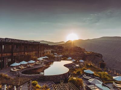 Luxuriöses Hotel mit Pool auf Felsen, Sonnenuntergang über Berglandschaft am Horizont.
