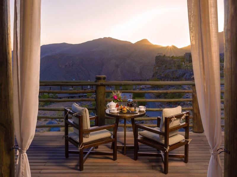 Balcony with table and four chairs overlooking mountains at sunset.
