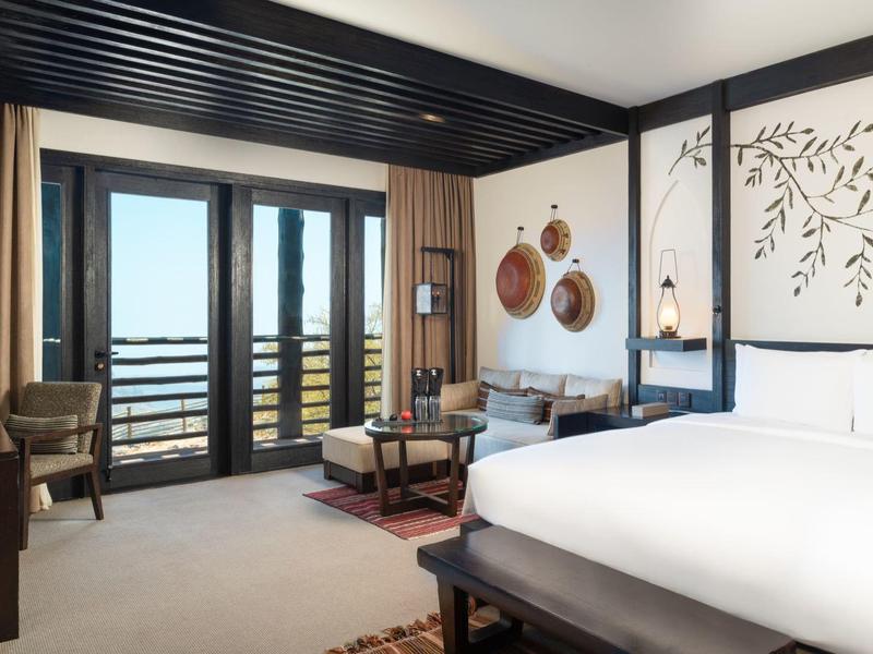 Modern hotel room with double bed, seating area, large windows, and balcony with sea view.