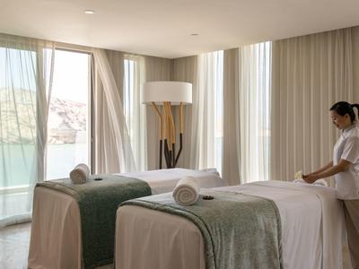 Lichte spa kamer met twee massagetafels en een therapeut die een tafel klaarmaakt.