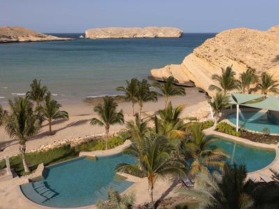 Strandlandschaft mit Palmen, zwei blauen Pools und felsiger Küste unter blauem Himmel.