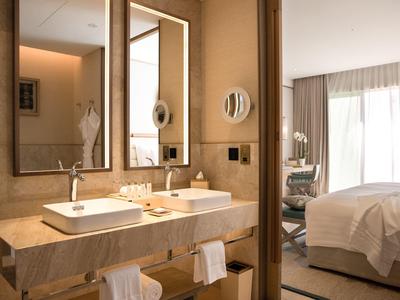 Moderne hotelbadkamer met dubbele wasbakken en grote spiegels naast een slaapkamer met natuurlijk licht.