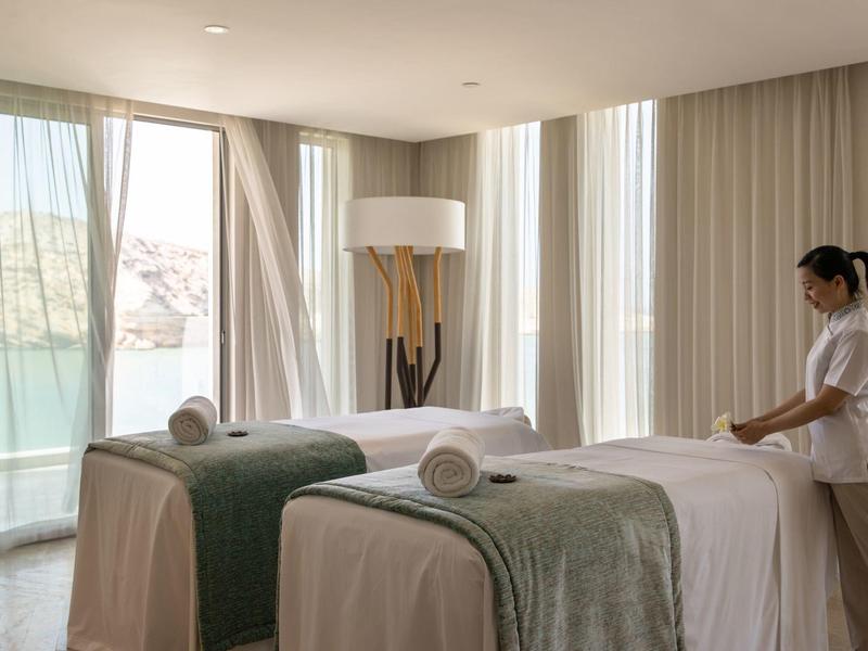 Lichte spa kamer met twee massagetafels en een therapeut die een tafel klaarmaakt.