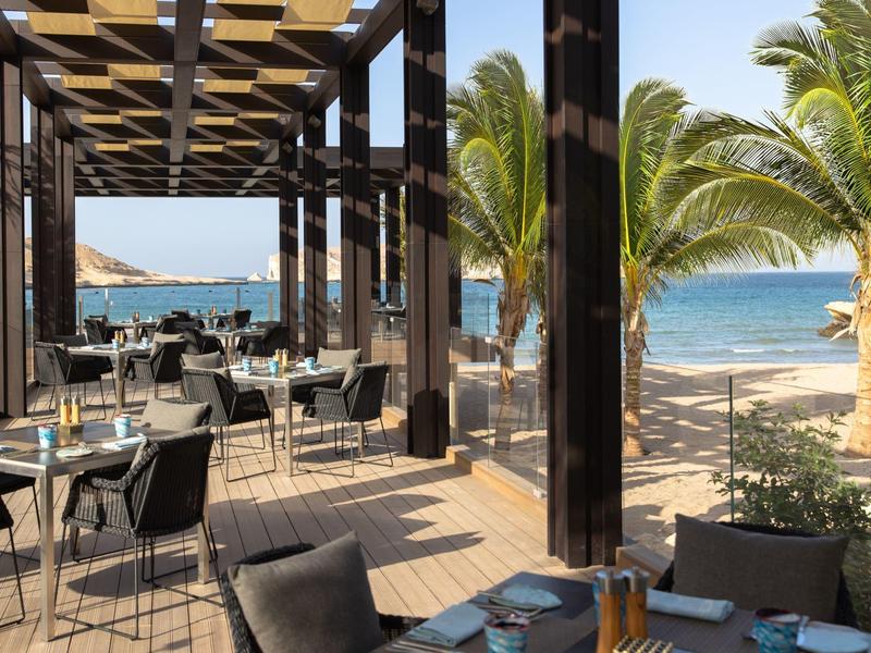 Buitenterras met tafels en stoelen aan het strand, uitzicht op zee en palmbomen.