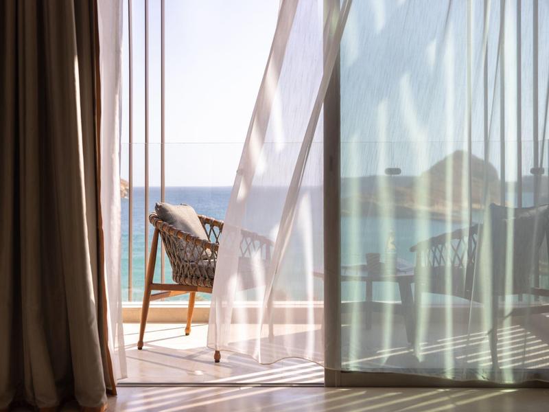 Open balkon deur met doorschijnende en donkere gordijnen, rotan stoel buiten met uitzicht op oceaan.