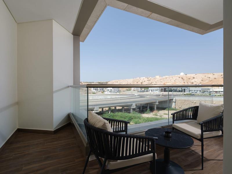 Balcony with two chairs, small table, glass railing overlooking buildings and hills.
