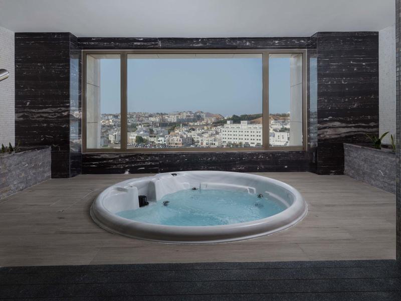 Indoor area with round whirlpool bathtub in front of large window with city view.
