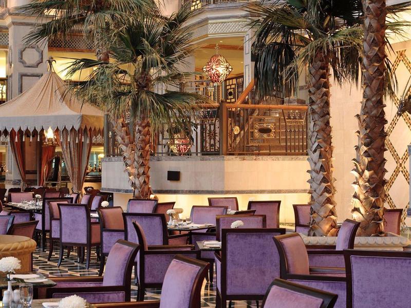 Salón de hotel elegante con sillas moradas, mesas de cristal y palmeras interiores.