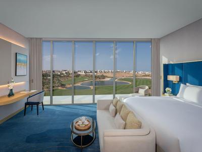 Modernes Hotelzimmer mit großem Bett, Sofa und Blick auf einen Golfplatz durch bodentiefe Fenster.