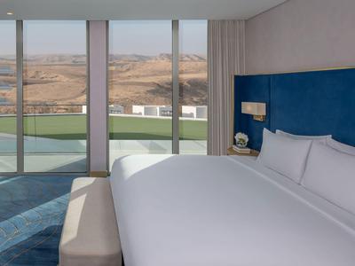 Modernes Hotelzimmer mit großem Bett, blauer Wand und Blick auf eine Wüstenlandschaft.