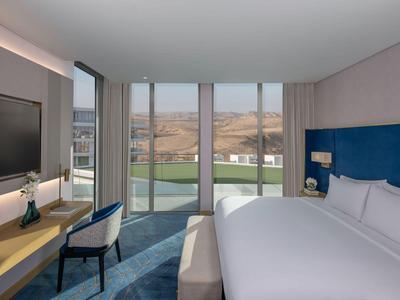 Modernes Hotelzimmer mit großem Bett, Schreibtisch, Stuhl und Blick auf Landschaft durch bodentiefe Fenster.