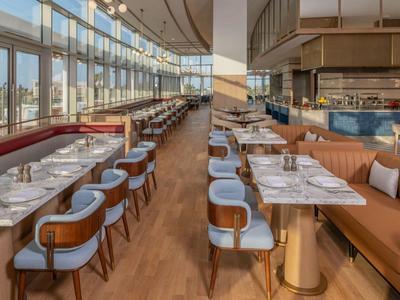 Modernes Restaurant mit großen Fenstern, Holzboden, Tischen mit weißen Marmorplatten und bequemen Stühlen.