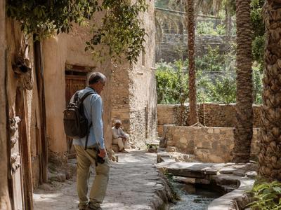 Ein Mann mit Rucksack steht auf einem Weg neben einem Wasserlauf und Palmen in einer Oasenumgebung.