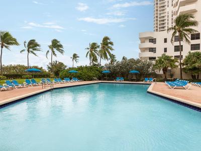 Duży hotelowy basen z niebieskimi leżakami i palmami w tle.