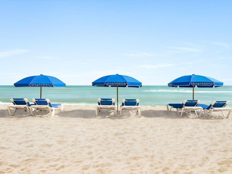 Trzy niebieskie parasole i leżaki na piaszczystej plaży nad spokojnym morzem.