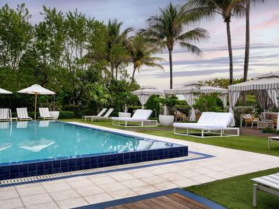 Schwimmbad mit blauen Fliesen, weißen Liegen, Sonnenschirmen, Palmen und grünem Garten bei Sonnenuntergang.