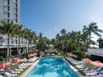 Großer Außenpool mit Liegestühlen und roten Schirmen, umgeben von Palmen und hohem Hotelgebäude.