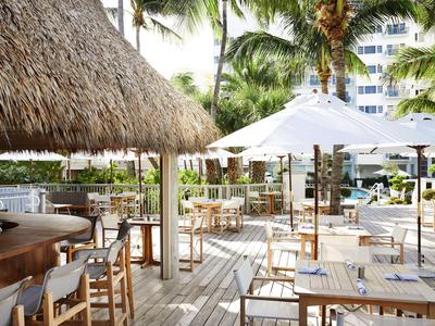 Außenbereich einer Hotelbar mit Holzmöbeln, Strohdach und Sonnenschirmen zwischen Palmen.