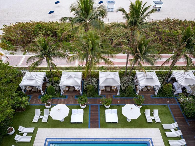 Blick auf einen rechteckigen Pool mit Liegestühlen, Palmen, Strand und Meer im Hintergrund.