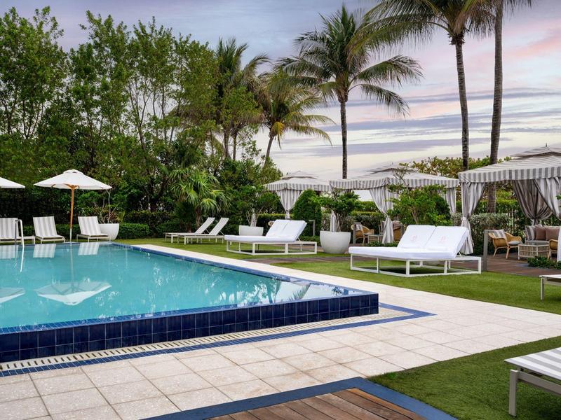 Schwimmbad mit blauen Fliesen, weißen Liegen, Sonnenschirmen, Palmen und grünem Garten bei Sonnenuntergang.