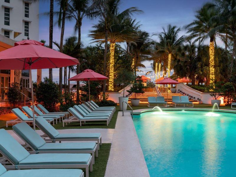 Beleuchteter Pool mit Liegestühlen und roten Sonnenschirmen, umgeben von Palmen und Hotels.