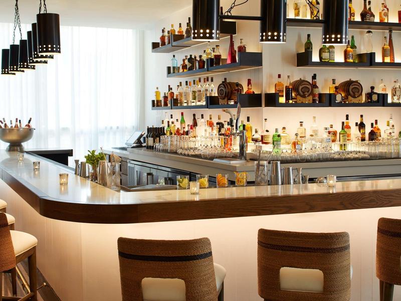 Moderne Bar mit hölzernen Barhockern, beleuchteten Regalen voller Flaschen und dunklen Hängeleuchten.