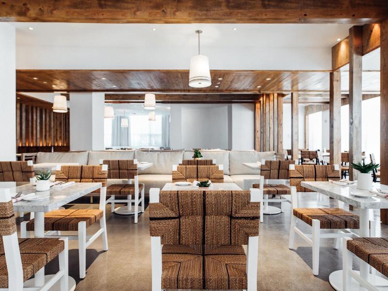 Modernes Restaurant mit weißen Tischen, Holzbalken und beigen, gepolsterten Stühlen in hellem Ambiente.