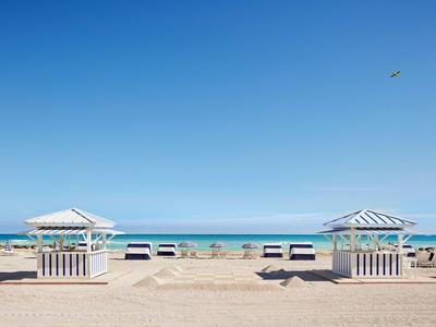 Sandstrand mit weißen Holzschirmen, blauer Himmel und ruhigem Meer im Hintergrund.
