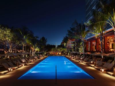 Beleuchteter rechteckiger Pool in tropischer Hotelanlage bei Nacht mit Palmen und Liegestühlen.