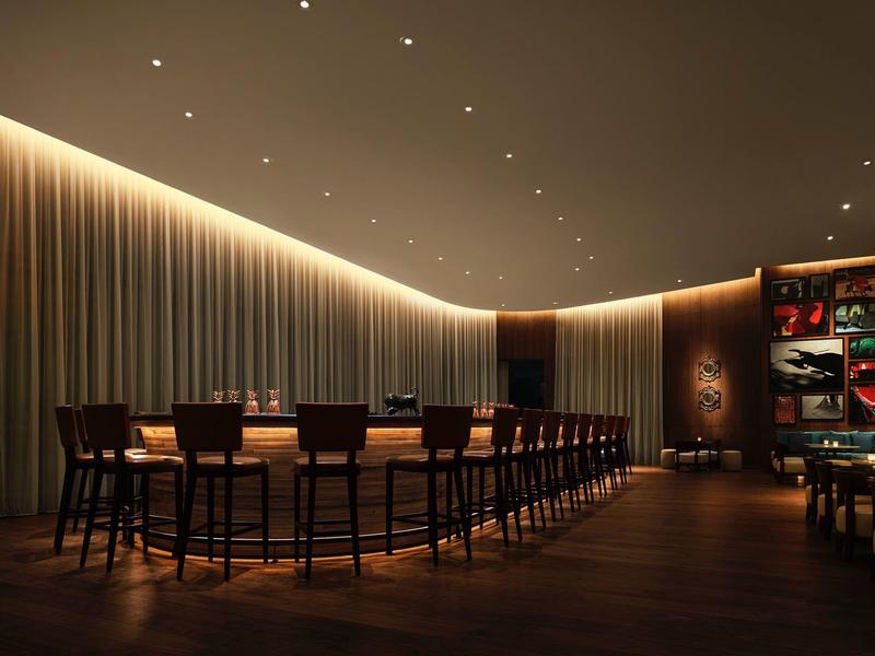 Moderner Barbereich mit langem Holztresen, hohen Stühlen und indirekter Beleuchtung an der Decke.